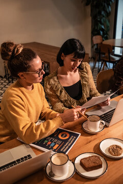 Two women reviewing document in cafe workspace with laptop