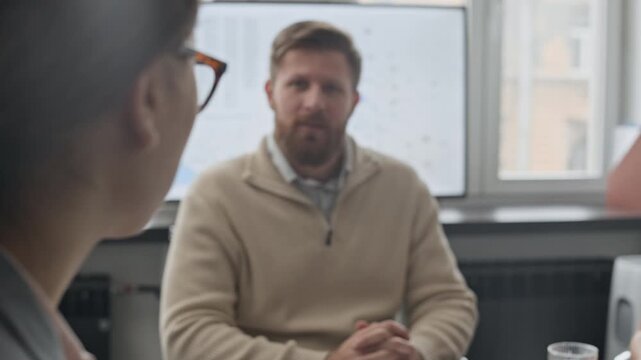 Over shoulder view of anonymous young woman sitting at desk, looking at her colleagues and boss while explaining her business idea during offline meeting in office