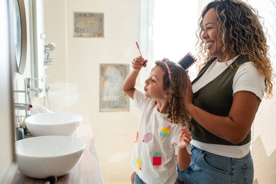 Mother helping daughter brush curly hair in bright bathroom