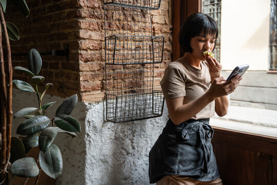 Young bakery worker tasting pastry while checking phone