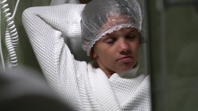 A girl looks at herself in a mirror in a hotel bathroom and adjusts the clear shower cap on her hair. The teenager is performing her morning or evening body care routine.