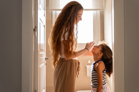 Mother helping daughter brush teeth in sunny bathroom