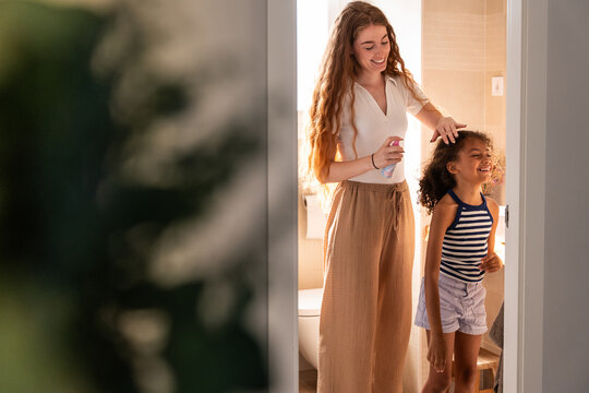 Mother sprays daughter hair in sunlit bathroom at home