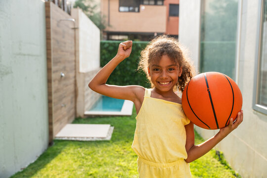 Smiling girl holding basketball and flexing in sunny backyard portrait