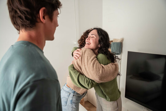 Happy couple hugging at home while friend watches nearby