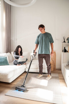 Young man vacuuming rug while woman works on laptop