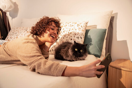 Smiling woman taking selfie with fluffy black cat on bed