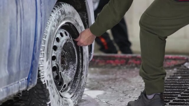 A blue car is being washed at a self-service car wash, fresh shampoo foam running down the body and wheels. The mechanic unscrews the tire pressure cap. High quality FullHD footage