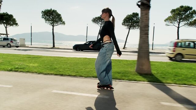 Gimbal full length tracking shot of young woman in wide jeans skating on sunny coastal road. Pine and palm trees, green lawn, sports car, sandy beach, sea and mountains behind. Mediterranean active 