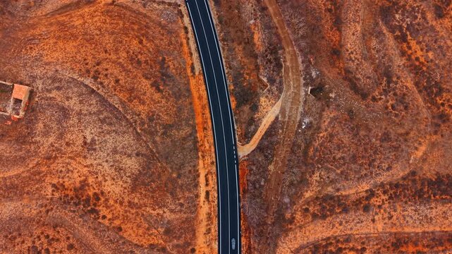 Vertical drone view of asphalt road and stream. Vertical drone view of straight black road beside dry red terrain, narrow stream, and small car.