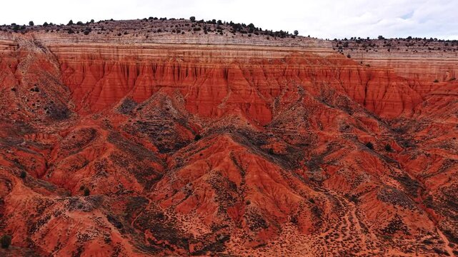 Red badlands slopes below layered cliffs in Spain. Oblique drone view of red eroded hills below horizontal pale strata and sparse vegetation.