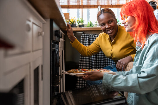 Two women smile while taking pizza out of oven at home