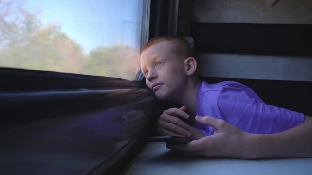 A red-haired teenager puts his phone down on the table and looks dreamily out the train window while sitting in his compartment during a journey. High quality FullHD footage
