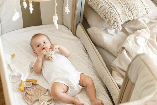 Cute little baby lying in crib with sweet toy and boho mobile. Bedtime. Adorable infant portrait in bassinet, space for text. Childhood and childcare.