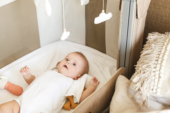 Cute little baby lying in crib with sweet toy and boho mobile. Bedtime. Adorable infant portrait in bassinet and smiling, space for text. Childhood and childcare.
