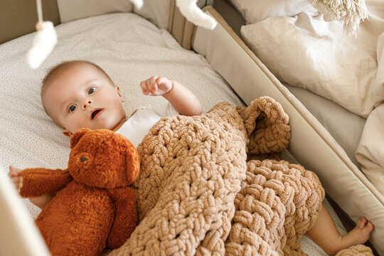 Adorable baby lying in crib with cute little toy teddy under knitted blanket. Time for sleep. Sweet infant portrait in boho bassinet top view. Childhood