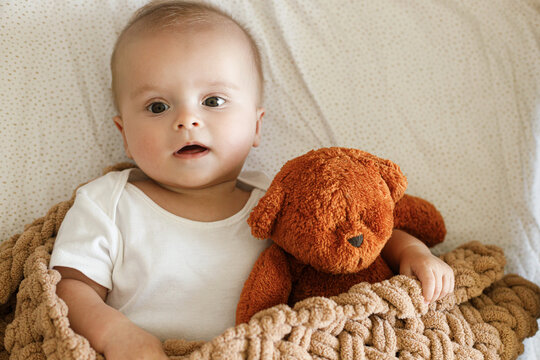 Adorable baby lying in crib with cute little toy teddy under knitted blanket. Time for sleep. Sweet infant portrait in boho bassinet top view. Childhood
