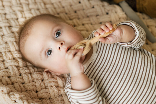 Cute baby lying with wooden teether on knitted blanket. Adorable infant portrait holding wooden toy, space for text. Childhood and childcare