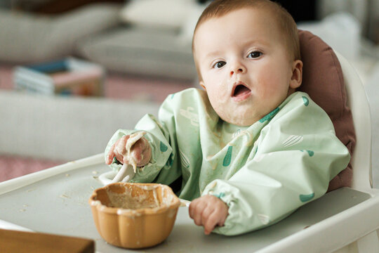 Cute little baby try to eat solid food in child chair. Complementary food. Adorable happy messy and funny infant portrait in porridge. Feeding after baby 6 months