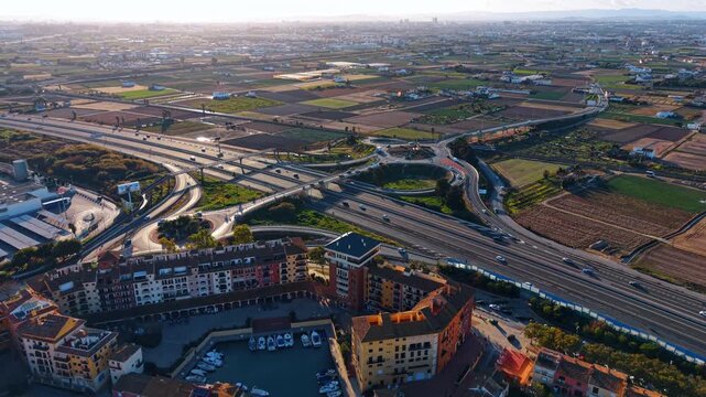 Wide aerial highway interchange near farmland and marina. Wide aerial view of highway interchange near fields, buildings, marina basin, and distant city skyline.