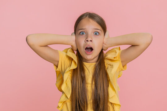 Young child girl covers ears with both hands, opening eyes wide in sudden fear panic caused by a loud sound or noise. School kid isolated on pink background, reacting emotionally with dramatic gesture