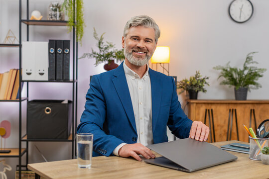Middle-aged businessman at home office closes laptop gazing camera proud after finishing project. Freelancer guy at table smiles satisfied reflecting achievement today confident posture up.