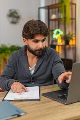 Fototapeta premium Indian man at home reviews paper reports compares numbers with laptop writes notes focused. Arabian guy at desk checks rows and figures on sheet reads data finds match does careful analysis calm mood