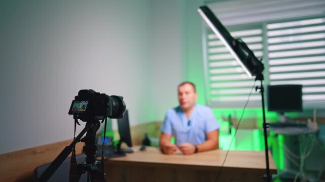 Clinic Vlog Setup With Doctor Camera And Light. Wide clinic setup with camera on tripod, LED tube light, desk, and doctor blurred in background.