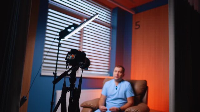 Behind Scenes Doctor Vlog Recording In Clinic Room. Behind scenes view of camera filming doctor in clinic room with orange wall and window blinds.