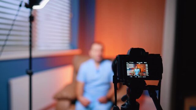 Doctor Recording Vlog In Orange Lit Clinic Room. Camera records doctor in blue scrubs in orange lit clinic room with LED light and chair.