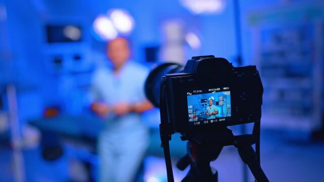 Close Camera Films Doctor Vlog In Blue Clinic. Close camera screen shows doctor in scrubs, blurred blue clinic background, tripod and LED light.