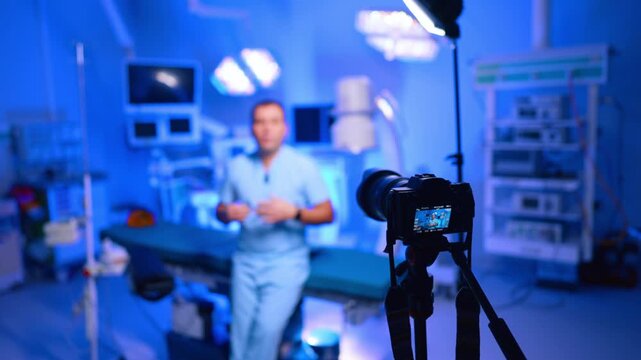 Video Camera Captures Doctor Vlog In Operating Room. Video camera screen shows doctor in scrubs, blurred operating room, blue lighting and medical devices.