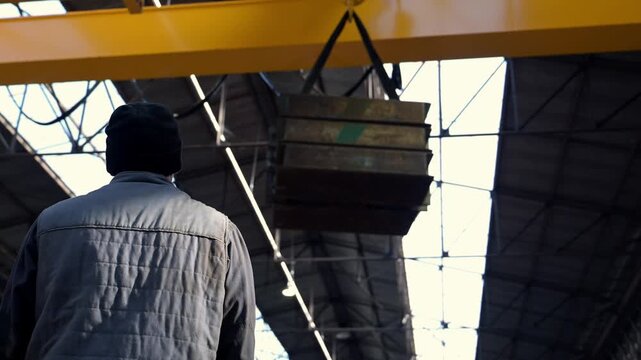 Industrial worker supervising an overhead crane lifting heavy steel plates in slow motion, showcasing precision, safety and large-scale factory operations.