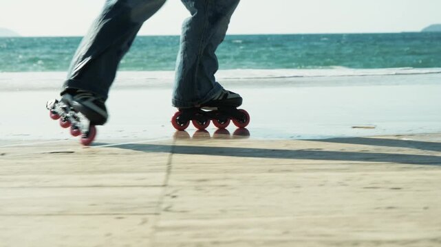 Close-up of roller skates moving along wet beach surface near sea, filmed with stabilizer in smooth parallel tracking shot. Ocean waves and sunlight reflections create dynamic summer lifestyle scene.