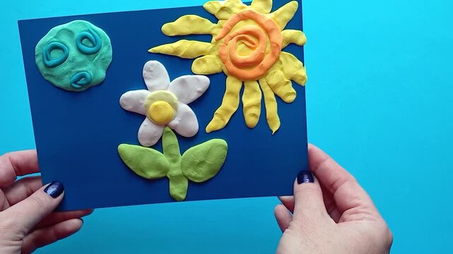 Hands represent a finished clay modeling product (flower, cloud, sun) on a blue background. Accelerated