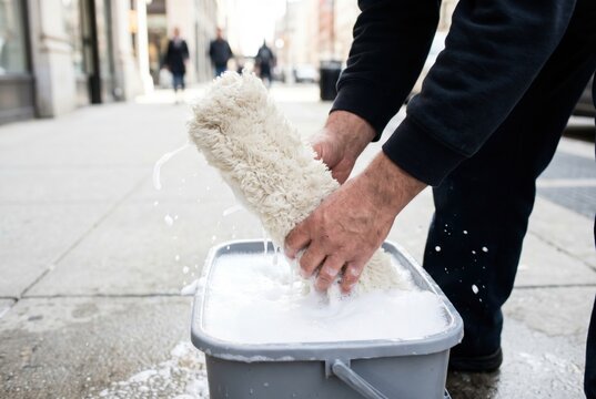 Window cleaner hands forcefully wringing out a wet soapy fleece applicator tool into a grey bucket on an urban sidewalk