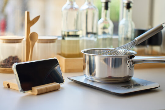Smartphone stands beside a saucepan with a whisk on a kitchen scale