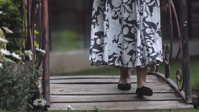 A woman walks toward the camera across a small garden bridge featuring wooden planks and metal handrails. She is dressed in a long floral skirt that remains elegant and subtle.
