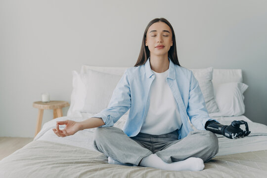 Young woman with prosthetic arm meditating on bed in bedroom at home