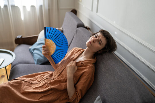 Sad woman with painful expression trying to cool down using paper fan in apartment without air conditioning, exhausted female waving hand fan sitting on sofa in living room, summer heatwave attack.
