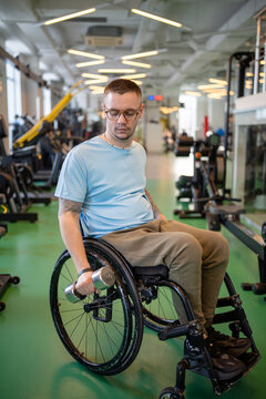 Man in wheelchair working out at gym on upper body and arms strength using dumbbell to build strong biceps muscles. Disabled male athlete with spinal injury do weight lifting training at sports center