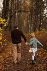 Fototapeta premium Back View Father And Daughter Walking Hand In Hand Down Leafy Trail Through Autumn Woods, Peaceful Reflective Scene With Golden Bushes, Slow Pace And Warm Seasonal Tones Evoking Journey