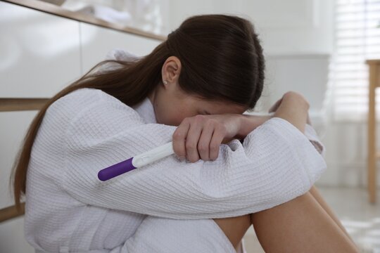 Frustrated woman in bathrobe with pregnancy test sitting near chest of drawers in bathroom