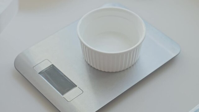 Hand adds baking soda from spoon into small bowl on digital kitchen scale. Concept of ingredient measuring and baking preparation