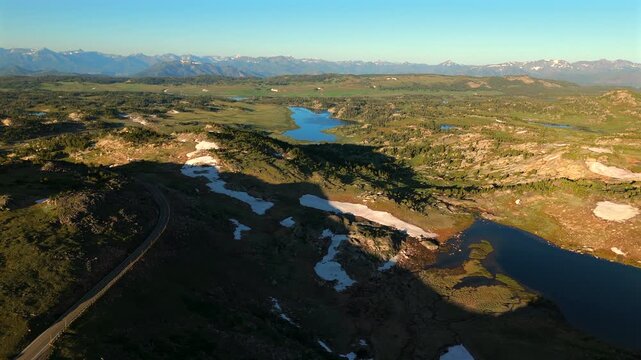 Beartooth Highway in Montana curves through stunning high elevation terrain with alpine lakes and melting snow patches. Distant snow-capped mountains frame the scenic wilderness landscape.