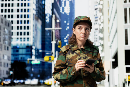 Using smartphone, city downtown and army soldier reading mission debriefing details from command center. Sending text messages back home, mobile phone to and woman wearing camouflage uniform