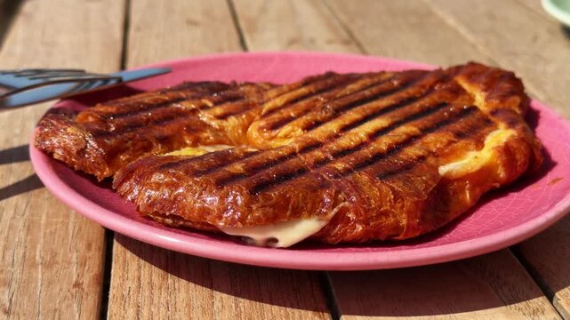 Grilled croissant planchado sandwich with ham and melted cheese on pink plate, woman girl cutting traditional breakfast pastry with knife and fork, crispy golden brunch food on wooden table, close up