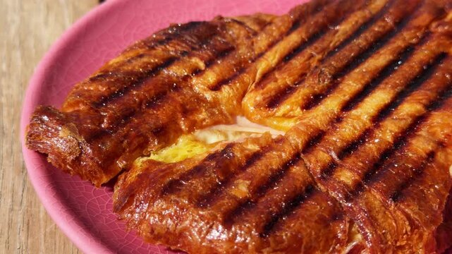 Grilled croissant planchado sandwich with ham and melted cheese on pink plate, woman girl cutting traditional breakfast pastry with knife and fork, crispy golden brunch food on wooden table, close up
