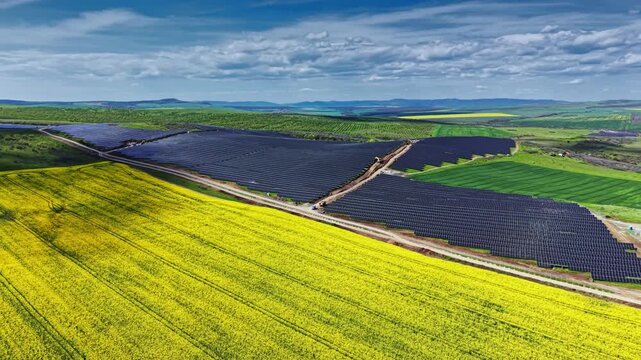 Workers are installing solar panels in a large field. The ground is flat and stretches for miles. Green hills are visible in the background. Clouds are in the sky.