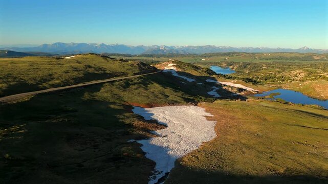 Aerial drone footage of Beartooth Highway winding through expansive alpine meadows during golden hour, featuring melting snow patches, pristine blue mountain lakes.